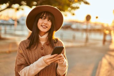 Kışlık şapkalı esmer kadın gün batımında şehrin dışında akıllı telefon kullanarak gülümsüyor.