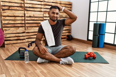 Spor salonundaki antrenman minderinde oturan genç Hintli adam gülümseyerek saçlara dokunuyor, el hareketi yapıyor, çekici ve modaya uygun poz veriyor. 