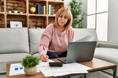 Orta yaşlı sarışın kadın evdeki kanepede oturarak tele-çalışmaya odaklanmış..