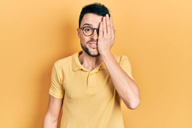 Gündelik kıyafetler ve gözlük takan genç İspanyol bir adam bir gözünü eliyle kapatıyor, yüzünde kendine güvenen bir gülümseme ve sürpriz bir duygu var.. 