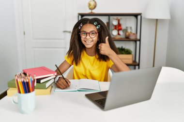 Young african american girl doing homework at home doing happy thumbs up gesture with hand. approving expression looking at the camera showing success. 