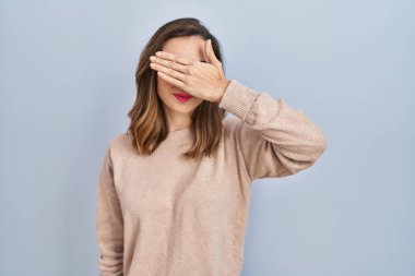Young woman standing over isolated background covering eyes with hand, looking serious and sad. sightless, hiding and rejection concept 