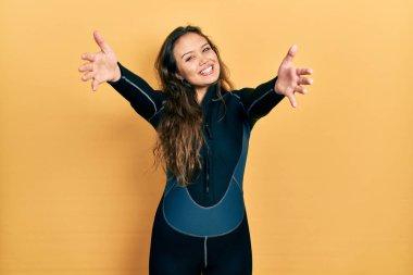 Genç İspanyol kız dalgıç neoprene üniforması giyiyor kameraya bakıyor ve sarılmak için kollarını açmış gülümsüyor. Mutluluğu kucaklayan neşeli bir ifade.. 