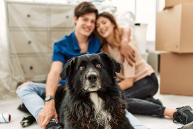 Genç beyaz çift mutlu bir şekilde gülümsüyor. Yeni evlerinde yerde köpekleriyle oturuyorlar..