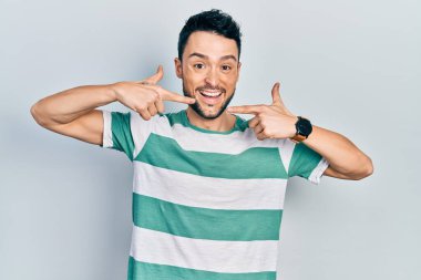 Günlük kıyafetler giyen, neşeli gülümseyen, parmaklarıyla ve ağzıyla işaret eden İspanyol genç bir adam. diş sağlığı kavramı. 