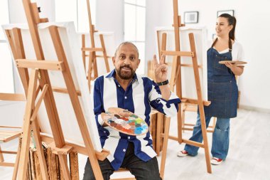 Sanat stüdyosundaki kıdemli sanatçı kameraya bakıp gülümseyerek parmaklarıyla zafer işareti yapıyor. İki numara.. 