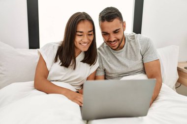 Yatak odasında dizüstü bilgisayar kullanarak mutlu gülümseyen genç Latin çift.