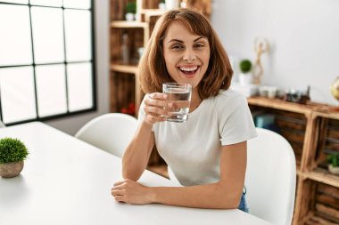 Beyaz kadın su içiyor. Evdeki masada oturuyor.