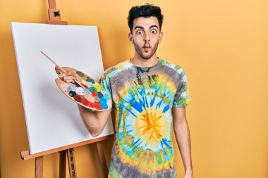 İspanyol asıllı genç adam paletle resim çiziyor, resim sehpası yapıyor, dudaklarıyla balık suratı yapıyor, çılgınca ve komik hareketler yapıyor. Komik ifade. 