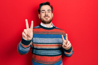 Sakallı, zarif yün süveter giyen yakışıklı beyaz adam kameraya bakıp zafer işareti yapıyor. İki numara.. 