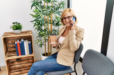 Orta yaşlı, sarışın, gülümseyen, kendine güvenen, bekleme odasında akıllı telefondan konuşan bir kadın.