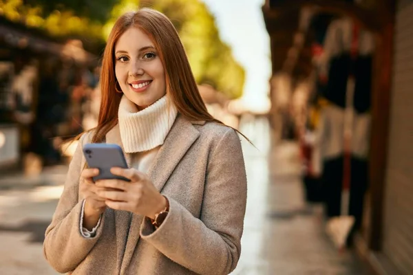Genç kızıl saçlı kız şehirde akıllı telefon kullanarak mutlu bir şekilde gülümsüyor..