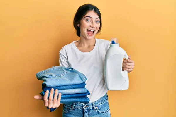 年轻的高加索女人一边洗衣服一边拿着洗涤剂瓶子和折叠的牛仔裤 一边笑着大声笑着 因为这是个可笑的疯狂笑话 — 图库照片
