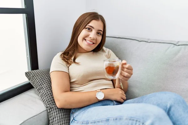 年轻的惊慌失措的女人带着自信的笑容在家里喝茶 — 图库照片