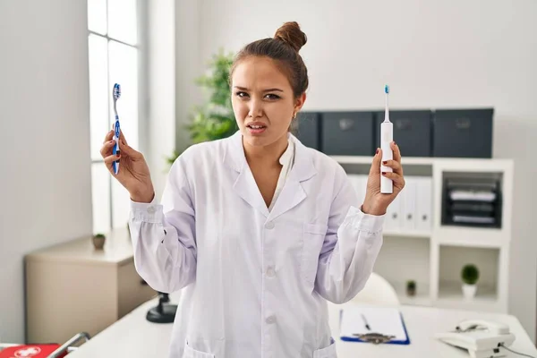 Young Hispanic Dentist Woman Holding Ordinary Toothbrush Electric Toothbrush Clueless — Stock Photo, Image