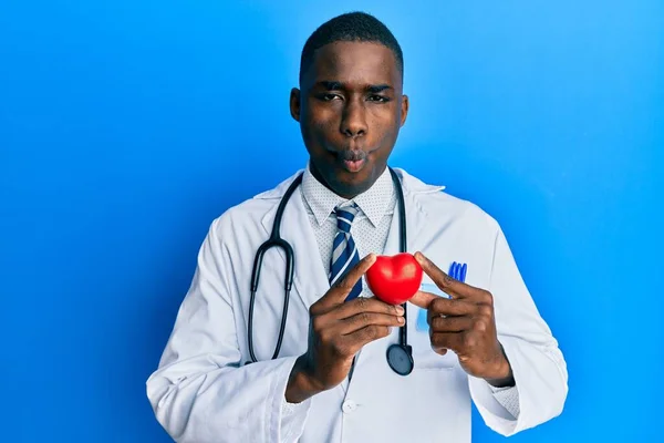 Young african american man wearing doctor uniform holding heart making fish face with mouth and squinting eyes, crazy and comical.