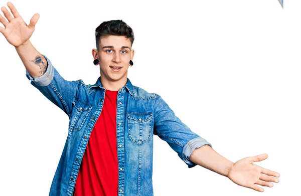 Young caucasian boy with ears dilation wearing casual denim jacket looking at the camera smiling with open arms for hug. cheerful expression embracing happiness. 