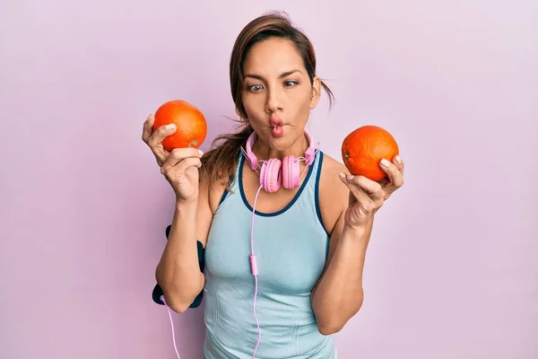 若いラテン語の女性は ヘッドフォンを使用して ジムの服を着て 口と目を細めて魚の顔を作るオレンジを保持し クレイジーでコミカルな — ストック写真
