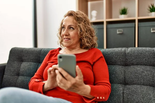 Orta yaşlı beyaz kadın akıllı telefon kullanıyor. Evdeki kanepede oturuyor..