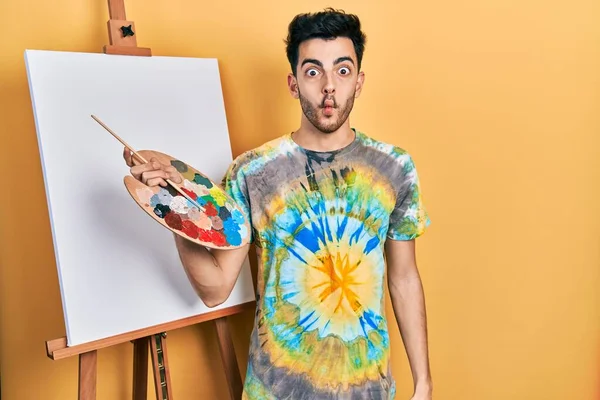 İspanyol asıllı genç adam paletle resim çiziyor, resim sehpası yapıyor, dudaklarıyla balık suratı yapıyor, çılgınca ve komik hareketler yapıyor. Komik ifade. 