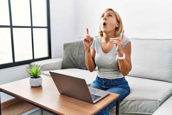 Блондинка средних лет, использующая ноутбук дома удивлена и удивлена, глядя вверх и указывая пальцами и поднятыми руками. 