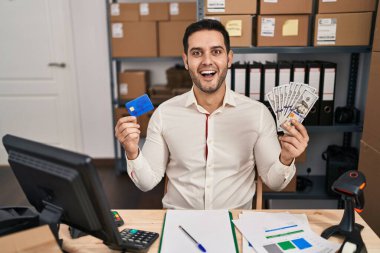 Sakallı genç İspanyol bir adam küçük işletme ecommerce çalışıyor. Elinde banknotlar ve kredi kartı var. Çılgınca kutlama yapıyor ve başarıyı haykırıyor.. 
