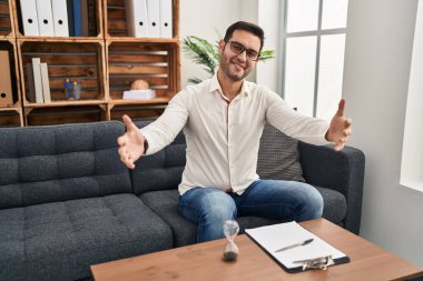 Danışma ofisinde çalışan sakallı genç İspanyol adam kameraya sarılıp gülümsüyor. Mutluluğu kucaklayan neşeli bir ifade.. 