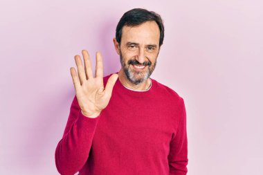 Orta yaşlı İspanyol bir adam, günlük kıyafetler giyip beş numarayı gösterip işaret ederken kendinden emin ve mutlu bir şekilde gülümsüyor.. 