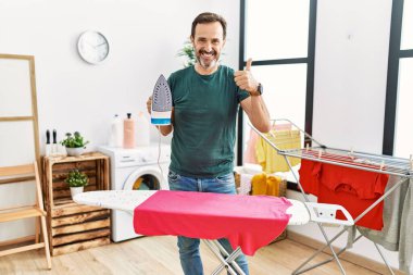 Evde sakallı, kıyafetlerini ütüleyen mutlu ve pozitif gülümseyen, başparmağı mükemmel ve onay işareti yapan orta yaşlı bir adam. 