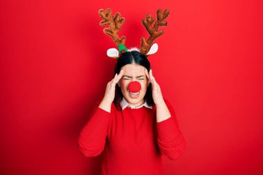 Geyik Noel şapkası takan genç İspanyol kadın ve eli başının üstünde kırmızı burun, stres yüzünden baş ağrısı. migren ağrısı. 