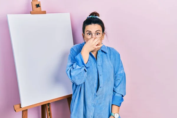 画家のイーゼルのそばに立つ若いヒスパニック系の女性は 鼻の上に指で息を保持し 臭いと嫌な 耐え難い匂いを嗅ぐ立っている 臭いが悪い — ストック写真