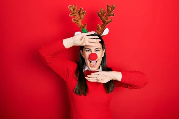 鹿のクリスマスの帽子をかぶっている若いヒスパニック系の女性と赤い鼻は陽気に顔を示す手で竹を覗かせて笑っています びっくりして興奮しました — ストック写真