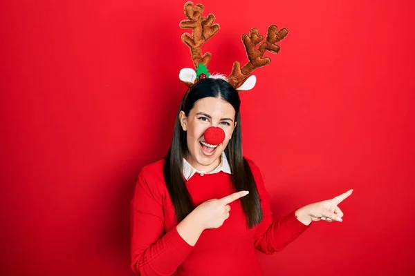若いですヒスパニック女性身に着けています鹿のクリスマスの帽子と赤い鼻笑顔とカメラを見てポインティングで二つの手と指で側面 — ストック写真