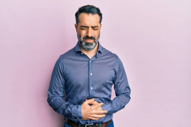 Orta yaşlı, sakallı ve gri saçlı bir adam midesi üstünde iş kıyafetleri giyiyor çünkü hazımsızlık, acı verici bir hastalık ve kendini iyi hissetmiyor. Ağrı kavramı. 
