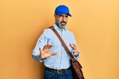 Orta yaşlı, sakallı ve gri saçlı kurye şapkalı bir adam tiksinti ifadesi takındı ve tiksinti reaksiyonu yüzünden tiksinti ifadesi takındı. Eller havada. 