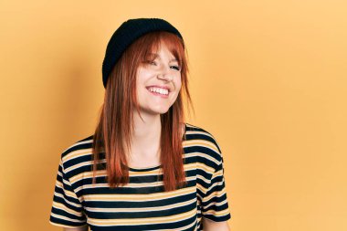 Kızıl saçlı, yün şapkalı, yüzünde gülümsemeyle yan yana bakan genç bir kadın, doğal bir ifade. Kendinden emin gülen. 