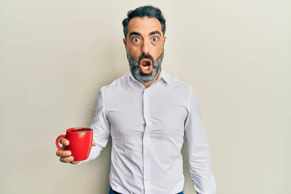 Middle age man with beard and grey hair drinking a cup coffee scared and amazed with open mouth for surprise, disbelief face