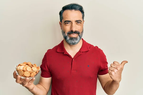Middle age man with beard and grey hair holding bowl with salty crackers biscuits pointing thumb up to the side smiling happy with open mouth