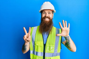 Uzun sakallı, güvenlik kaskı takan ve yansıtıcı ceketli kızıl saçlı adam yedinci parmaklarını gösterip gösterirken kendinden emin ve mutlu gülümsüyor.. 