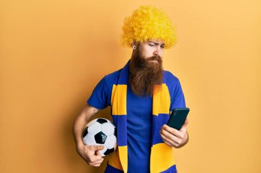 Kızıl saçlı, uzun sakallı futbol holiganı tezahürat yapan, elinde akıllı telefon tutan, yüzünde ciddi bir ifadeyle rahatlamış bir adam. Basit ve doğal bir şekilde kameraya bakıyorsun.. 