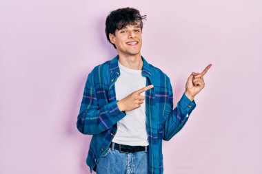 Yakışıklı hippi genç adam gündelik beyaz tişört ve klasik gömlek giyiyor gülümsüyor ve kameraya bakıyor iki eliyle ve parmaklarıyla yan tarafa bakıyor.. 