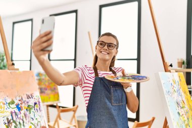 Gülümseyen genç bir kadın sanat stüdyosundaki akıllı telefondan özçekim yapıyor.