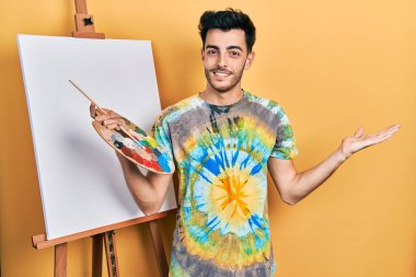 İspanyol asıllı genç adam paletle resim çiziyor resim sehpası ayakta gülümsüyor mutlu bir şekilde sunuyor ve avuç içi kameraya bakıyor.. 