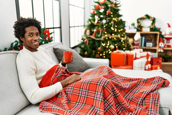 若いアフリカ系アメリカ人の男性は 幸せとクールな笑顔でクリスマスツリーでソファの上に座ってコーヒーを飲んでいます 運のいい人 — ストック写真