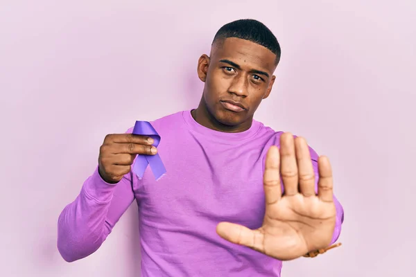 Young black man holding purple ribbon awareness with open hand doing stop sign with serious and confident expression, defense gesture