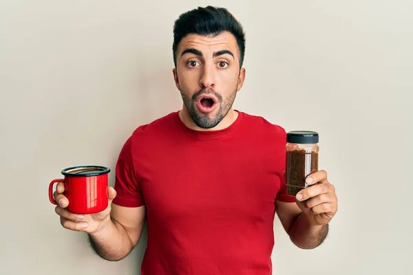 Young hispanic man holding cup of soluble coffee afraid and shocked with surprise and amazed expression, fear and excited face.