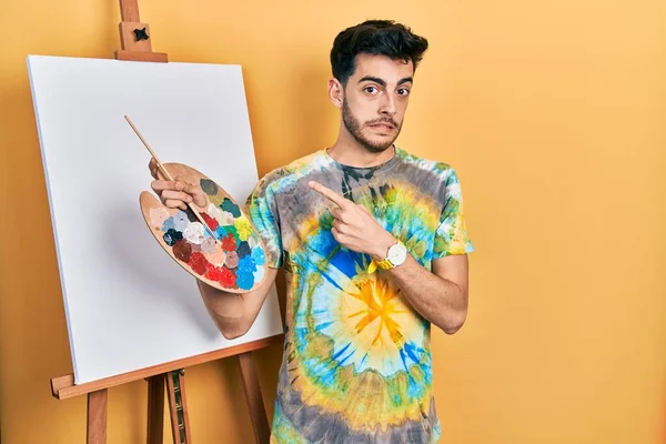 画家によってパレットで描かれた若いヒスパニック系の男イーゼルスタンドは 前指で心配し 緊張を脇に置きます 心配し 驚きの表情 — ストック写真