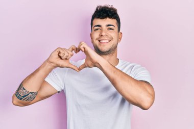 Gündelik beyaz tişört giyen, aşık gülümseyen, elleriyle kalp sembolü yapan genç İspanyol adam. romantik kavram. 