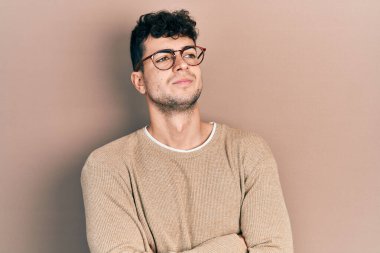 Gündelik kıyafetler ve gözlük takan genç İspanyol adam kollarını kavuşturmuş ve kendinden emin bir şekilde yan tarafa bakıyor. 