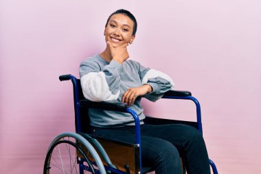 Tekerlekli sandalyede oturan güzel İspanyol kadın kameraya kendinden emin bir şekilde bakıyor kollarını kavuşturmuş ve çenesini kaldırmış gülümsüyor. Olumlu düşünmek. 